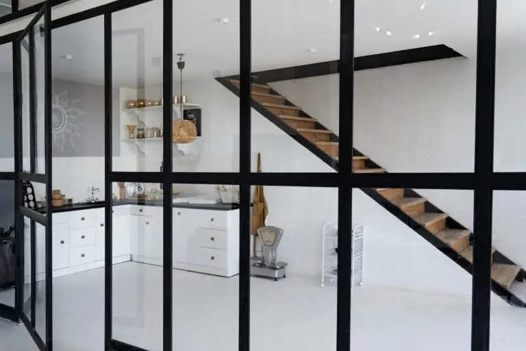 Verrière noire d'intérieur séparant une cuisine blanche, escalier en bois